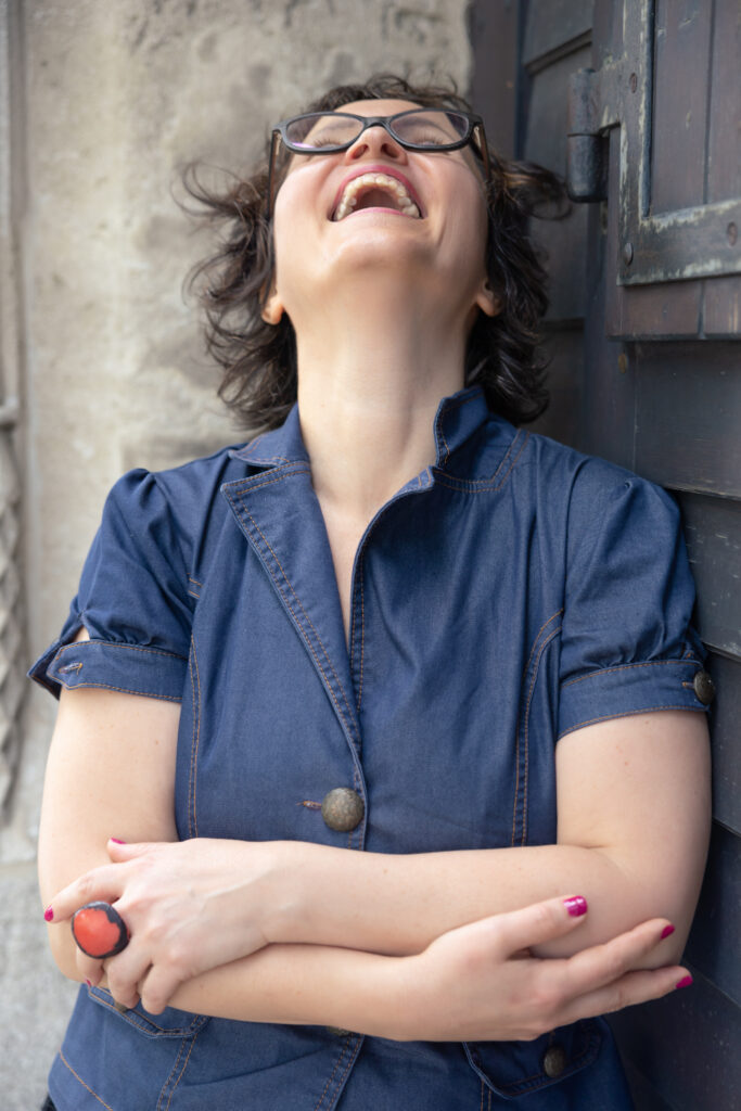 Edit Árokszállási mit Brille, vor Holztür, lacht, blaues Outfit, roter Ring